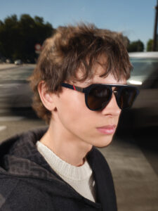 A young person with short, tousled hair wears large, dark sunglasses in an outdoor setting. The attire includes a dark jacket over a light, textured sweater. The background features a blurred street scene with passing vehicles and trees, under a clear blue sky, capturing a sense of motion and a crisp, bright day.