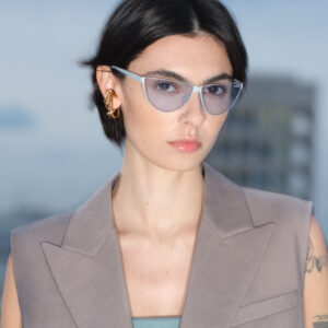 A person with short dark hair wears stylish, translucent blue glasses and a taupe, sleeveless blazer. Gold hoop earrings accentuate their ears. The background is softly blurred, hinting at a urban environment with a building and sky. Their expression is poised and confident, complementing the overall sophisticated and modern aesthetic.