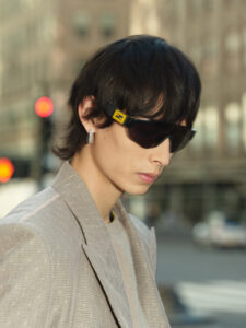 A person stands on a city street, wearing dark sunglasses and a light-colored, textured jacket over a matching top. Their dark hair falls around the ears, complemented by a sleek, modern earring. The background shows an urban setting with blurred buildings and subtle traffic lights, softly out of focus, adding depth to the scene.