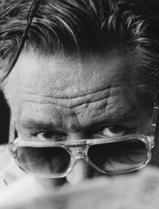 A close-up of a man's face features his intense gaze peering over the rim of transparent sunglasses. Deep, expressive lines mark his forehead, and his hair is styled back, showing strands of gray. This monochromatic view captures a serious and contemplative expression, with shadow and light accentuating the texture of his skin and facial details.