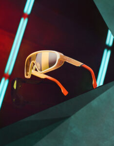 A pair of futuristic sunglasses with a sleek, glossy design features an amber-shaded frame and reflective lenses. The frames have vibrant orange detailing on the temples. Neon lights cast soft blue and red glows, enhancing the modern aesthetic against a dark, angular background. The combination creates an atmosphere of cutting-edge style and dynamic contrast.