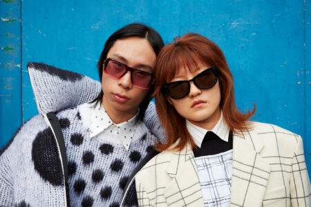 Two individuals are posed against a bright blue backdrop, exuding a retro vibe with their bold sunglasses. The left person wears a polka-dot sweater over a patterned shirt, while the right person sports a geometric-patterned jacket with a high-collared shirt. Their hairstyles are sleek and distinctive, adding to the striking contrast with the vivid background.