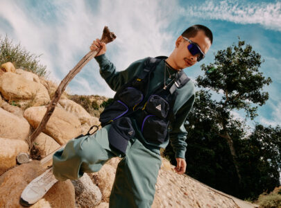 A person wearing sunglasses, layered clothing, and a utility vest with pockets stands on rocky terrain. They hold a stick while looking downwards, surrounded by stones and sparse vegetation. In the background, a tree and a bright blue sky with wispy clouds create a sense of adventure and exploration in the natural landscape.