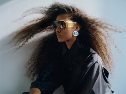 The composition features a person with voluminous, windswept hair, styled in loose curls. They wear large, round sunglasses with a golden frame, paired with oversized, circular earrings. The attire is a dark, collared shirt, contributing to a sleek and modern look. Soft lighting creates gentle shadows, highlighting the subject's profile and the dynamic flow of their hair.