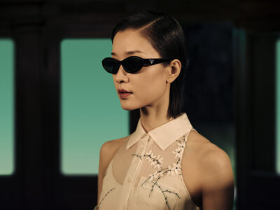 A person with short, sleek hair and wearing small, black sunglasses looks confidently to the side. They are dressed in a light, sleeveless blouse with floral embroidery, set against a softly blurred background of teal and dark tones. The lighting highlights the smooth texture of their skin and the elegant lines of the outfit’s design.