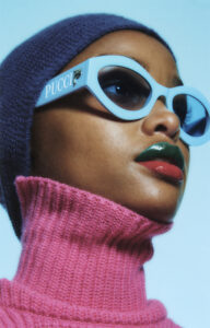 A person gazes upward, exuding confidence and style. They wear bold, oversized sunglasses with a striking, light blue frame. Their lips are painted in vivid green and red shades, contrasting with a bright pink, textured turtleneck sweater. A dark beanie covers their head, adding a touch of warmth to the ensemble against a light blue background.