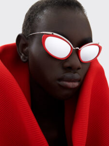 A person wears bold, reflective red-framed eyewear, exuding a futuristic vibe. The individual is draped in a striking red garment featuring a textured design. The overall aesthetic is both modern and avant-garde, with a minimalist, neutral background that emphasizes the vibrant contrast of red against the person's skin and accessories.