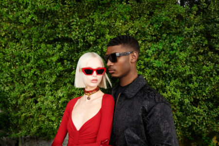 A stylish duo stands closely against a lush green backdrop. The person on the left wears a red outfit, highlighted by bold red sunglasses and a decorative choker. Their companion sports a dark, textured jacket paired with chic black sunglasses. The scene suggests elegance and modern fashion set in a serene, natural environment.
