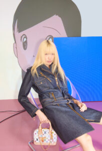 A person with long blonde hair wears a dark denim outfit with gold zippers, sitting on a transparent chair. They hold a small, colorful, patterned handbag. The setting features a background with an animated character's face, using vibrant colors like pink and blue, suggesting a modern, stylish atmosphere with a blend of fashion and pop culture elements.