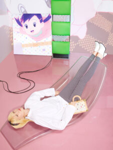 A person with blonde hair lies on a transparent chaise lounge against a pink background. They wear a white shirt and striped pants, holding a small, stylish bag. Next to them, a colorful animated character is displayed on a screen, accompanied by a modern, geometric green structure. Black cables trail across the floor, adding a dynamic element.