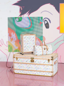 A colorful, artistic setup displays three stylish pieces of luggage featuring a vibrant pattern. The largest piece, a trunk, serves as the base for a smaller rolling suitcase and a chic duffle bag. Behind them, an eye-catching backdrop of playful, cartoonish art adds a dynamic feel, complementing the luxurious, travel-themed arrangement on a glossy surface.