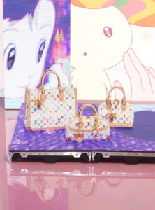 Three stylish handbags with a colorful pattern combining logos and symbols sit on a reflective pink surface. The bags feature light leather handles and gold hardware, including padlocks and key tags. A backdrop displays dreamy animated characters in soft pastel tones, enhancing the playful yet fashionable setting. Each bag varies slightly in shape and size.