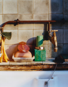 A rustic kitchen sink area features a weathered brass faucet against aged tiles. On the ledge, colorful stacked soaps rest in a dish beside a green scrub pad. Nearby, a watch rests atop a solid green block. Hanging towels add warmth to the scene, capturing a lived-in, vintage atmosphere with natural lighting highlighting the textures.