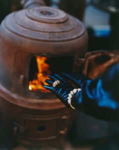 A pair of hands wearing black gloves and gold accessories is positioned in front of a rustic cast iron stove. The stove door is open, revealing a warm, glowing fire inside. The surrounding atmosphere suggests a blend of industrial and cozy elements, contrasted by the sleek gloves and detailed jewelry adorning the hands.