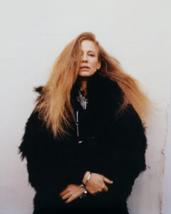 A person with long, light brown hair appears contemplative against a plain background. They wear a luxurious black fur coat, layered necklaces, and a bracelet, exuding a chic, edgy style. Their arms are crossed, partly hidden by the coat, while their intense gaze is directed forward. The overall mood is one of sophistication and mystery.