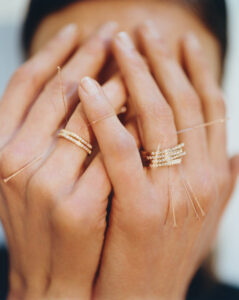 Two hands are held together, fingers interlaced over a blurred face. The skin has a soft, natural tone. Delicate rings adorn the fingers, with thin bands and tiny gemstones. Golden threads hang from some of the rings, adding an intricate detail. The background is softly out of focus, enhancing the hands' prominence and elegance.