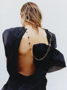 The composition features a person with wet, tousled hair facing away, revealing an exposed back. They wear a loose, deep black garment that falls off one shoulder, exposing a delicate chain adorned with decorative elements draping down the spine. The overall aesthetic conveys a sense of effortless elegance and minimalistic style, with a subdued and neutral backdrop.
