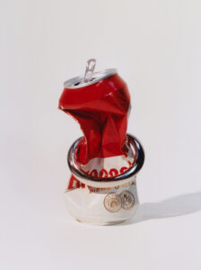 A crumpled red and white aluminum can is artistically pierced by a shiny metal ring. The can, partially squashed, stands upright on a plain surface. The juxtaposition of the crushed texture and smooth, reflective ring creates an intriguing visual contrast. Light and shadow interplay highlights the can's creases and the ring's sleek surface, adding depth.