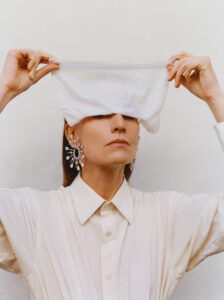 A person stands against a neutral background, wearing a light-colored, collared shirt. Large, ornate earrings add a glamorous touch. Their arms are raised, holding a white cloth over their eyes, creating a sense of elegance and mystery. The expression is serene and poised, emphasizing a unique blend of fashion and artistic expression.