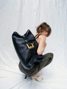 A person with short brown hair crouches, wearing a woven, olive-green skirt and light-colored, open-toe heels. They hold a large black bag with a prominent gold buckle. The setting is minimalist with a softly crumpled white backdrop, creating a contemporary and stylish atmosphere, emphasizing the blend of fashion and elegance in the pose and accessories.