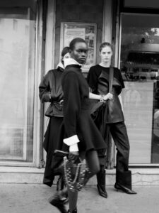 Three individuals stand in an urban setting, dressed in stylish, monochromatic outfits. The first person, in the foreground, carries a purse with chain details, exuding confidence. The others stand slightly behind, exhibiting a relaxed yet poised stance. The scene takes place in front of a shop window and a textured exterior wall, creating a modern, fashion-forward atmosphere.
