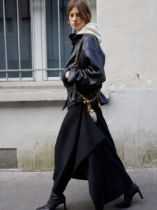 A person with short hair walks confidently along a sidewalk, wearing a dark leather jacket over a light hoodie. They sport wide-legged black pants and stylish black ankle boots. A gold chain accessory with a small furry detail hangs from the outfit. The background features a barred window and a textured white wall.