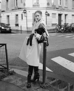 A person leans against a pole at a street intersection. They wear a long coat, boots, and hold a bag. The setting features an urban background with bicycles parked along the sidewalk and a building with shuttered windows. The scene is monochromatic, capturing a classic, stylish moment with an emphasis on urban fashion and elegance.