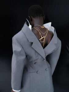 A person with short dark hair wears a gray double-breasted suit, styled open at the back to reveal a white shirt beneath. Two gold chains, featuring elaborate key-like pendants, drape across the back. The background is a solid, dark color, highlighting the striking contrast and unique fashion ensemble. The overall atmosphere is avant-garde and artistic.
