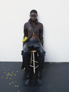 A person sits confidently on a stool, dressed in a dark leather jacket and matching pants. They hold yellow flowers in one hand, resting on their lap. Scattered petals lie on the dark floor. The backdrop is a simple, minimalist white wall, emphasizing the sleek and stylish attire, creating a striking and fashion-forward appearance.
