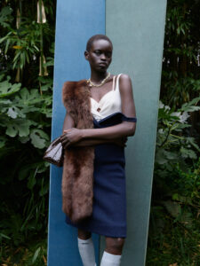 A person is standing in front of a lush, green backdrop, framed by tall foliage and vertical panels in shades of blue. They are wearing a cream top with a blue skirt, accessorized with a fur stole and a brown handbag. The look is completed with white knee-high socks and a delicate necklace, combining elegance with natural elements.