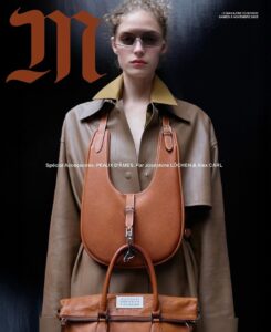 A person with tousled, curly hair wears a tan leather coat with a pointed collar. They sport sleek, futuristic sunglasses. Two leather handbags, one small and another larger, drape over their shoulders, complementing the outfit. The background is a dark, solid color, enhancing the chic and modern aesthetic of the ensemble and accessories.