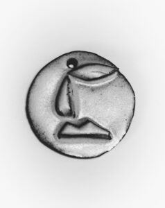 A circular, textured metallic object featuring a minimalist face design with simple lines and cutouts. The visage is abstract, with prominent facial features suggested through smooth indentations, creating a modern, artistic impression. The piece has a small hole near the top, possibly for hanging or attaching. The overall feel is both industrial and artistic.