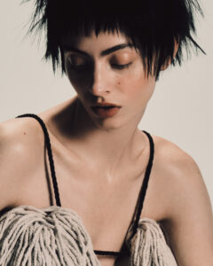 A person with short, dark, tousled hair appears in a thoughtful pose, head slightly tilted, and eyes looking down. The individual wears a strappy garment made of textured material, resembling wool or yarn, with a soft shadow cast on their skin. The overall tone is warm and earthy, highlighting a serene and introspective mood.