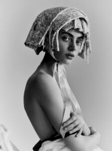 A person gazes with an intense expression, wearing an intricately textured headpiece tied with ribbons around the chin. Their arms are crossed, partially draped in white fabric, conveying a sense of vulnerability and strength. The monochrome palette accentuates the fine details of the headpiece, the soft shadows on the skin, and the overall serene yet contemplative mood.