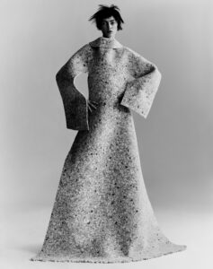 A monochrome portrayal features an individual with short, tousled hair wearing a long, textured garment with wide sleeves. The attire resembles a large, intricate fabric with a speckled pattern, draping gracefully to the floor. The person stands confidently, hands on hips, exuding an avant-garde, sculptural elegance against a neutral backdrop, capturing a bold fashion statement.
