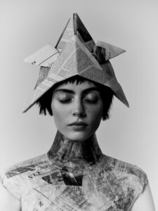 A young woman wears a hat crafted from folded newspaper, with a serene expression and eyes gently closed. Her attire integrates newspaper textures, mimicking clothing with a patchwork of overlapping pages. The monochromatic style enhances the creative, avant-garde atmosphere, emphasizing her peaceful demeanor and the artistic composition of paper elements blending with her form.