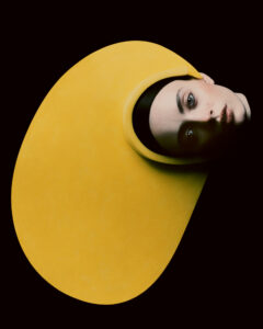 A person is depicted with a striking, oversized yellow hat covering a significant portion of the surrounding area. The background is dark, accentuating their pale face and blue eyes. The minimalistic composition creates a dramatic contrast, blending fashion and art in a modern, avant-garde style. The expression is neutral, adding an enigmatic quality to the scene.