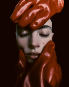 A serene face is encased by three pairs of shiny red gloves. Closed eyes suggest tranquility or introspection, while the gloves create a striking contrast with the soft features. The positioning of the hands defines the composition, adding a dramatic visual element. The color palette consists mostly of reds and muted skin tones, set against a dark background.