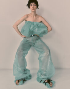 A person with short dark hair stands confidently against a plain background, wearing an unconventional outfit made entirely from transparent bubble wrap. The ensemble includes a voluminous top and wide-legged pants, accentuated by matching footwear. The pose and clothing evoke a sense of avant-garde fashion, blending elements of whimsy and innovation in design with a playful twist.