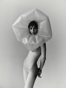 A person stands against a plain background, adorned with a unique headpiece crafted from bubble wrap, giving a dramatic, avant-garde look. Their short hair complements the minimalist style, and the bare torso is slightly turned, creating dynamic lines. The overall composition is striking, combining fashion-forward creativity with a touch of surrealism.