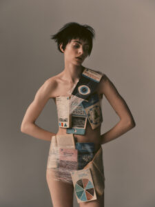 A person with short, dark hair stands against a plain background. Various colorful books and pages are strategically taped across their body, creating a unique and artistic collage. The individual gazes downward with a contemplative expression. The overall mood is thought-provoking and avant-garde, blending elements of literature and fashion in an unconventional manner.