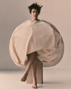 A person with short, tousled dark hair sits on a wooden block against a neutral background. They wear an oversized, spherical garment that envelops the upper body, leaving the arms concealed. The legs extend gracefully below the spherical dress, with bare feet resting lightly on the floor. The overall composition is minimalist and avant-garde.