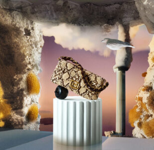 A beautifully patterned bag with a snake-like texture and a prominent gold accent rests on a cylindrical pedestal. The background features a surreal landscape with stone formations and a column-like structure. The scene includes a bird gliding gracefully, enhancing the whimsical atmosphere with the soft hues of a sunset creating a dream-like setting.