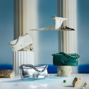 A surreal scene features three elegant purses in varying colors: white, metallic silver, and deep green. Each purse has a distinctive chain strap and is artfully placed among classical columns and abstract elements like a rocky surface and fragmented pedestal. The background displays a serene gradient, creating a harmonious blend of art and fashion in a dreamy setting.