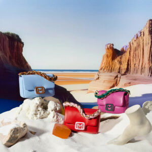 Three colorful bags are artistically placed against a backdrop of striking orange sandstone formations and a serene blue sky. The bags, in vibrant shades of blue, pink, and red, feature decorative braided handles. They are positioned on white, textured rocks, with sculptural elements adding to the composition. The setting exudes a sense of luxury and elegance.