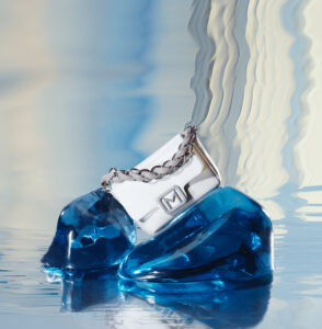 A sleek, silver-colored accessory with a chain handle is artfully positioned atop glossy, blue crystalline shapes. The background displays a gradient of soft, flowing hues, blending from blue to cream, creating a tranquil, reflective ambiance. The overall composition conveys elegance and modernity through the fusion of metallic elements and vibrant, translucent forms.