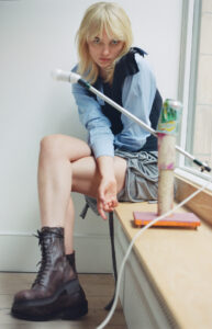 A blonde individual with straight hair sits on a window ledge, wearing a layered outfit including a blue shirt and tie. They sport brown lace-up boots and look directly ahead. A decorative object with a colorful can is nearby. The setting appears minimalist with light-colored walls and wooden flooring, creating an artistic, contemplative atmosphere.