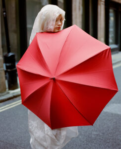 In an urban setting, an individual is partially concealed by a vivid red umbrella. They are wrapped in a translucent, patterned fabric, featuring delicate floral designs, with the material covering their head like a veil. The street is empty, with the background comprising tall buildings and a grey asphalt road, suggesting a quiet yet dynamic city scene.