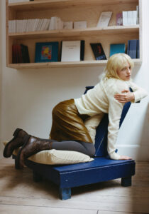 A person with blonde hair is positioned on a contemporary blue chair, resting on plush cushions. Dressed in a cozy cream sweater, brown skirt, and dark tights, they wear sturdy brown boots. The setting includes wooden flooring and a spacious wall shelf adorned with books and colorful covers, creating a relaxed, artistic atmosphere.