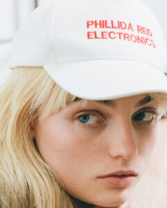 A close-up portrait showcases a person with long, blonde hair wearing a white cap adorned with stitched red text. The focus is on the individual's expressive blue eyes and subtle facial features. The light hits softly, highlighting the natural complexion and delicate skin texture, conveying a sense of understated style and casual elegance.